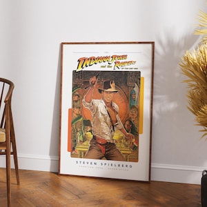 May include: A vintage movie poster for the 1981 film "Indiana Jones and the Raiders of the Lost Ark." The poster features a brown-haired man in a tan shirt and brown pants holding a whip. The poster is framed in a brown wood frame.