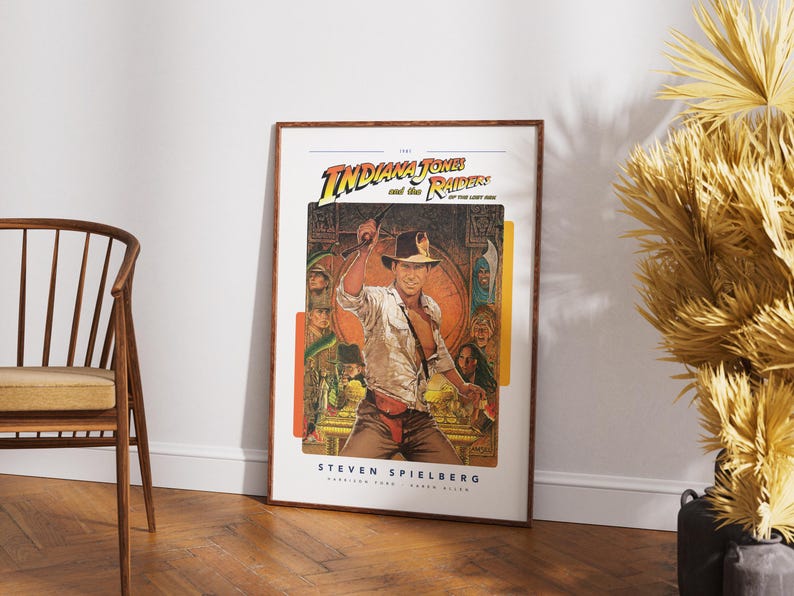 May include: A vintage movie poster for the 1981 film "Indiana Jones and the Raiders of the Lost Ark." The poster features a brown-haired man in a tan shirt and brown pants holding a whip. The poster is framed in a brown wood frame.