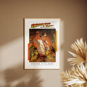 May include: A vintage movie poster for the film "Indiana Jones and the Raiders of the Lost Ark." The poster features a colorful illustration of Indiana Jones holding a whip and standing in front of a temple. The text "Steven Spielberg" is printed at the bottom of the poster.