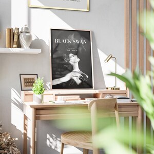 May include: A home office desk with a wooden top and legs. A black and white poster with the text "BLACK SWAN" and "NATALIE PORTMAN" is hanging on the wall above the desk. A small potted plant is on the desk, along with a lamp and a few books.