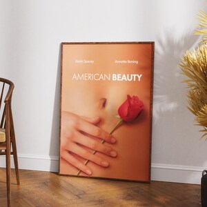 Op de afbeelding: Een ingelijste filmposter voor "American Beauty" met een close-up van een torso met een hand die een rode roos vasthoudt. De poster heeft de namen Kevin Spacey en Annette Bening bovenaan. De titel "AMERICAN BEAUTY" staat in grote witte letters.