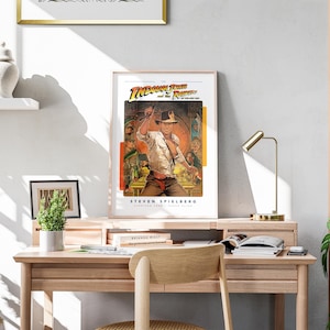 May include: A framed movie poster for the film "Indiana Jones and the Raiders of the Lost Ark" featuring a scene of Indiana Jones holding a whip and standing in front of a temple. The poster is on a desk with a lamp and a plant.