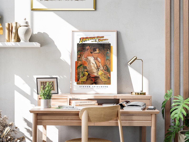 May include: A framed movie poster for the film "Indiana Jones and the Raiders of the Lost Ark" featuring a scene of Indiana Jones holding a whip and standing in front of a temple. The poster is on a desk with a lamp and a plant.