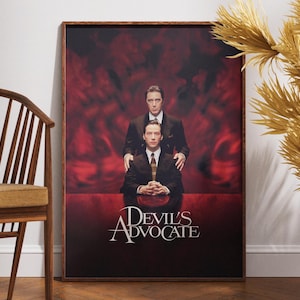 May include: A framed movie poster for "Devil's Advocate" featuring two men in suits against a red background. The title is displayed in elegant white lettering. The poster is in a dark wooden frame.