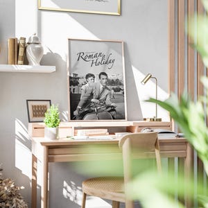 Może przedstawiać: Czarno-biały plakat z tekstem "Roman Holiday" na jasnym drewnianym biurku ze złotą lampą i doniczką z rośliną.