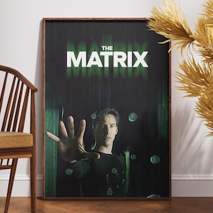 May include: A framed poster for the movie "The Matrix" featuring a man reaching out with his hand. The title is in white block letters against a green and black background. The poster is in a brown wooden frame.