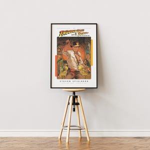 May include: A vintage movie poster for the film "Indiana Jones and the Raiders of the Lost Ark" featuring a color illustration of Indiana Jones holding a whip and standing in front of a golden idol. The poster is framed in black and is hanging on a white wall above a wooden stool.
