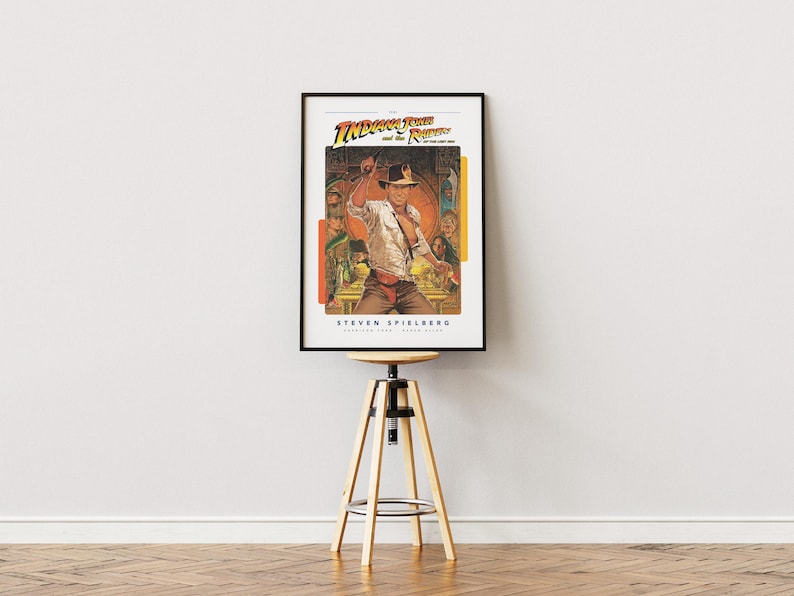 May include: A vintage movie poster for the film "Indiana Jones and the Raiders of the Lost Ark" featuring a color illustration of Indiana Jones holding a whip and standing in front of a golden idol. The poster is framed in black and is hanging on a white wall above a wooden stool.
