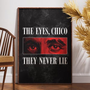 May include: Framed art print with the text "THE EYES, CHICO THEY NEVER LIE" in white on a black background. The artwork features a red illustration of eyes. A wooden chair and decorative plant are in the background.