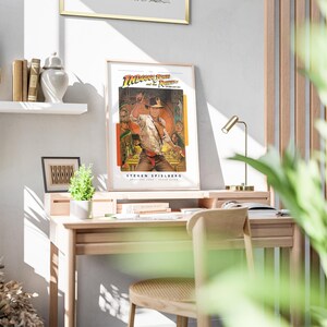 May include: A framed poster of the movie "Indiana Jones and the Raiders of the Lost Ark" featuring a scene of Indiana Jones with a whip in a jungle setting. The poster is hanging on a white wall above a wooden desk with a lamp and a plant.