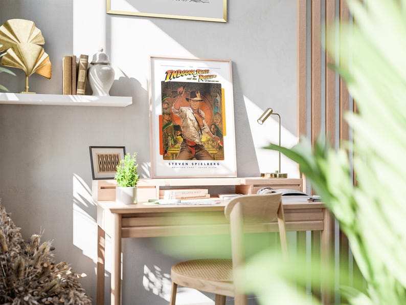 May include: A framed poster of the movie "Indiana Jones and the Raiders of the Lost Ark" featuring a scene of Indiana Jones with a whip in a jungle setting. The poster is hanging on a white wall above a wooden desk with a lamp and a plant.