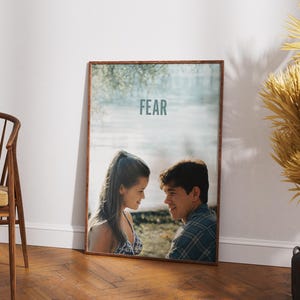 Op de afbeelding: Een ingelijste poster met een foto van een stel dat naar elkaar kijkt. De poster heeft een lichtblauwe achtergrond en het woord "FEAR" in witte tekst.