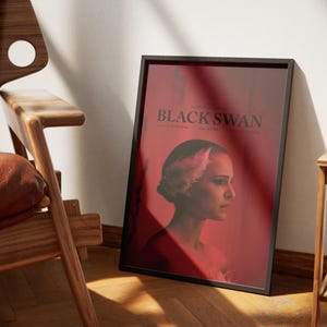 Puede incluir: Un p&oacute;ster enmarcado en negro con el t&iacute;tulo "Black Swan" en texto blanco. El p&oacute;ster presenta un primer plano de una mujer con un tocado de plumas blancas, mirando hacia la derecha. El fondo es un degradado rojo.