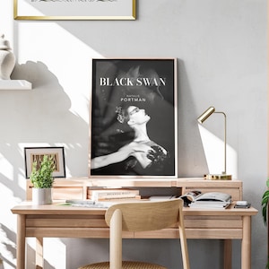 May include: A black and white poster of Natalie Portman as a black swan in the movie "Black Swan". The poster is framed and hanging on a wall above a wooden desk with a lamp and books.