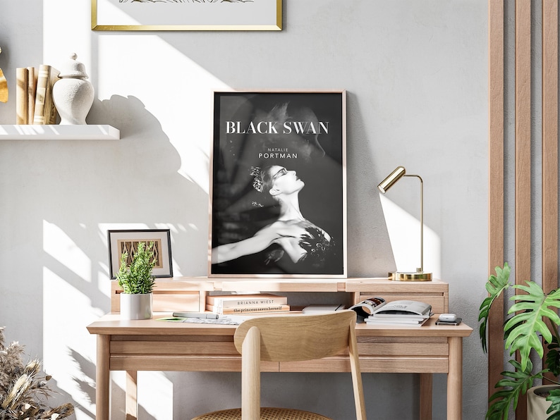 May include: A black and white poster of Natalie Portman as a black swan in the movie "Black Swan". The poster is framed and hanging on a wall above a wooden desk with a lamp and books.