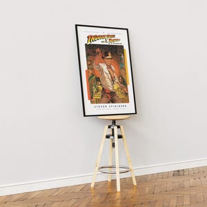 May include: A framed poster of the movie "Indiana Jones and the Raiders of the Lost Ark" featuring a scene of Indiana Jones holding a whip. The poster is on a wooden stool.