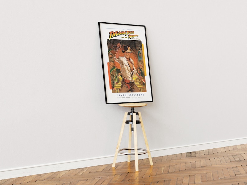 May include: A framed poster of the movie "Indiana Jones and the Raiders of the Lost Ark" featuring a scene of Indiana Jones holding a whip. The poster is on a wooden stool.