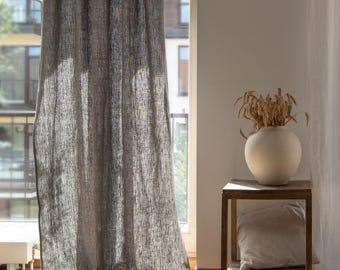 May include: A gray linen curtain hangs in front of a window, with a white curtain to the right. A small wooden side table holds a white vase with dried wheat and a beige pillow. The room is filled with natural light.