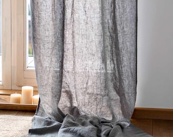 May include: A gray linen curtain hangs in front of a window, with natural light filtering through. The curtain is slightly bunched at the bottom, and three candles sit on a wooden windowsill. The curtain's texture is visible.