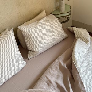 Può includere: Una scena di camera da letto con un letto con piumino e federe color rosa antico. Le federe e il copripiumino hanno un motivo a righe nei toni del bianco e del grigio. È visibile un piccolo comodino con un vaso di fiori secchi.