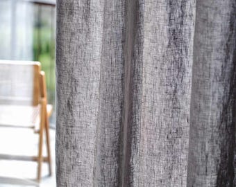 May include: Close-up of a gray linen curtain with vertical folds. The fabric has a textured appearance, with visible woven threads. A wooden chair and a balcony are visible in the background, suggesting a window treatment.