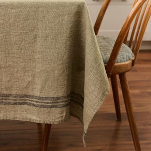 Peut inclure: Une nappe en lin dans les tons beige et jaune, avec un détail de rayures gris foncé près du bord inférieur. La nappe est drapée sur une table en bois, avec une chaise en bois visible en arrière-plan. Le tissu a un aspect texturé.