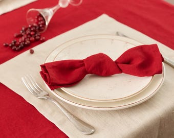 May include: A festive table setting with a red linen tablecloth and a beige linen placemat. A red linen napkin is tied in a bow on a white plate. A silver fork and a fallen glass with scattered cranberries complete the scene.