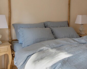 Puede incluir: Una cama con ropa de cama azul claro, que incluye una funda nórdica y fundas de almohada con volantes. La estructura de la cama es de bambú. Dos lámparas blancas con pantallas cuadradas a ambos lados de la cama.
