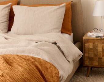 Könnte beinhalten: Ein Bett mit neutralfarbener Bettwäsche, darunter eine hellbeige Bettdecke und Kissen sowie eine rostfarbene Decke. Ein Nachttisch aus Holz mit einer Lampe und einer Brille steht neben dem Bett. Das Bettgestell ist mit einem passenden neutralen Stoff bezogen.