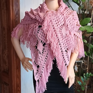 May include: A light pink, handmade crochet shawl with fringe. The shawl is draped over a mannequin. The crochet pattern features openwork designs. The shawl is a soft, feminine accessory.