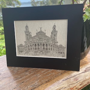 Puede incluir: Dibujo arquitectónico en blanco y negro enmarcado, que representa un gran edificio con torres y arcos. La obra de arte se presenta en un marco negro, sobre una superficie de madera, con un fondo natural.