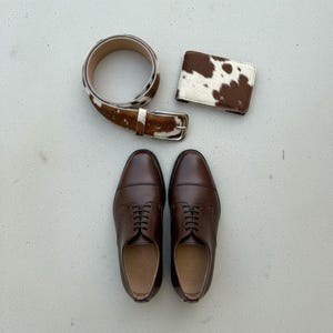 Puede incluir: Un conjunto de accesorios masculinos: un cintur&oacute;n de cuero marr&oacute;n con estampado de piel de vaca, una cartera a juego y un par de zapatos de vestir de cuero marr&oacute;n. Los zapatos son de estilo cl&aacute;sico.