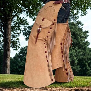 K&ouml;nnte beinhalten: Hellbraune Lederchaps mit Fransen und einer Tasche. Die Chaps haben ein dekoratives Design und werden mit einem G&uuml;rtel und einer Schnalle befestigt. Rote N&auml;hte akzentuieren die Kanten.