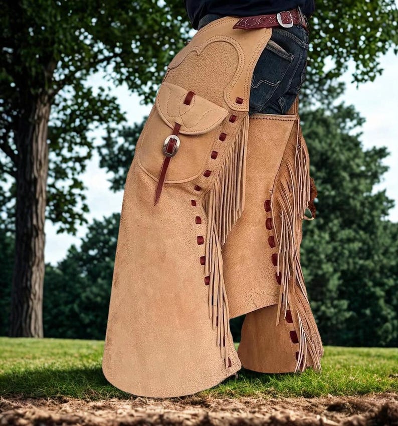 K&ouml;nnte beinhalten: Hellbraune Lederchaps mit Fransen und einer Tasche. Die Chaps haben ein dekoratives Design und werden mit einem G&uuml;rtel und einer Schnalle befestigt. Rote N&auml;hte akzentuieren die Kanten.