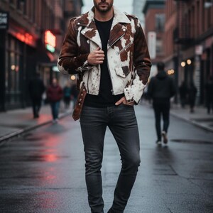 May include: A brown and white cowhide leather jacket with a black shirt and dark gray jeans. The jacket has a classic biker style with a notched collar, zipper closure, and a belt at the waist. The person is standing on a wet street.