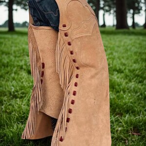 K&ouml;nnte beinhalten: Hellbraune Wildleder-Chaps mit dekorativen N&auml;hten und Fransen. Die Chaps haben rote Akzente und werden &uuml;ber einer blauen Jeans getragen. Das Bild ist vor dem Hintergrund von gr&uuml;nem Gras und B&auml;umen aufgenommen, was ein Western-Thema hervorruft.