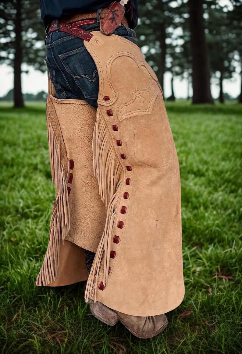 K&ouml;nnte beinhalten: Hellbraune Wildleder-Chaps mit dekorativen N&auml;hten und Fransen. Die Chaps haben rote Akzente und werden &uuml;ber einer blauen Jeans getragen. Das Bild ist vor dem Hintergrund von gr&uuml;nem Gras und B&auml;umen aufgenommen, was ein Western-Thema hervorruft.