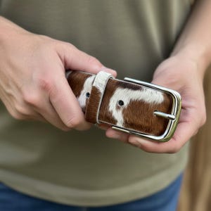 Puede incluir: Un cintur&oacute;n de piel de vaca marr&oacute;n y blanco con una hebilla plateada. El cintur&oacute;n se sostiene en las manos, mostrando el patr&oacute;n natural y la textura del cuero. La hebilla es rectangular con una sola hebilla.