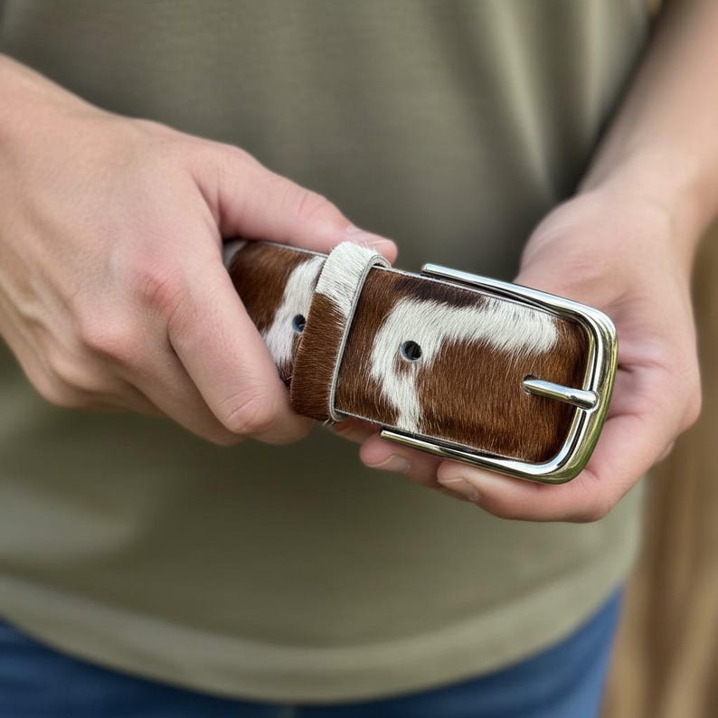 Puede incluir: Un cintur&oacute;n de piel de vaca marr&oacute;n y blanco con una hebilla plateada. El cintur&oacute;n se sostiene en las manos, mostrando el patr&oacute;n natural y la textura del cuero. La hebilla es rectangular con una sola hebilla.