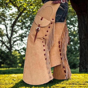 K&ouml;nnte beinhalten: Hellbraune Lederchaps mit Fransen und einer Tasche. Die Chaps haben dekorative N&auml;hte und einen Schnallenverschluss. Das Bild zeigt die Chaps &uuml;ber einer blauen Jeans, mit einem G&uuml;rtel an der Taille. Die Chaps sind zum Reiten gedacht.