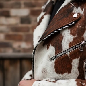 May include: A brown and white cowhide leather jacket with a silver zipper and a snap closure. The jacket has a classic biker style with a notched collar and a fitted silhouette. The jacket's pattern is a mix of brown and white patches.