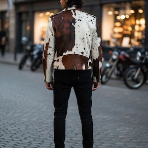 May include: A cowhide leather jacket with a brown and white spotted pattern. The jacket has a classic biker style with a notched collar, asymmetrical zipper, and shoulder epaulets. The person is wearing black jeans and black boots.