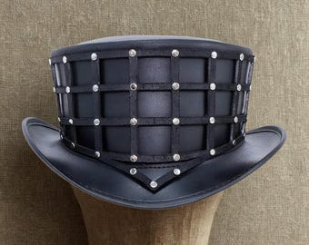 May include: A black top hat with a unique design. The hat features a grid-like pattern made of black straps and silver studs. The brim is wide and the hat is displayed on a mannequin head, showcasing its shape and style.