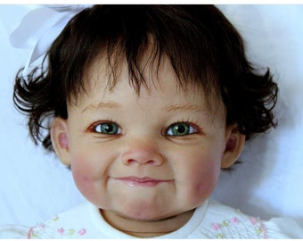Puede incluir: Primer plano de una muñeca bebé con cabello castaño y ojos verdes. La muñeca lleva un vestido blanco con flores rosas y blancas.