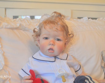 Puede incluir: Primer plano de una muñeca bebé con cabello rubio rizado que lleva una camisa blanca con cuello y ribete negro y un atuendo a rayas azul y blanco. La muñeca sostiene un juguete rojo.