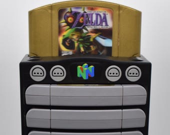Puede incluir: Un soporte de almacenamiento de cartuchos de videojuegos negro y gris con un cartucho dorado de "Zelda" insertado en la parte superior. La unidad tiene múltiples ranuras grises y la palabra "BAXA" está impresa en ella.
