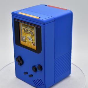 以下が含まれることがあります： 黒い画面の縁と黒いボタンが付いた青い携帯ゲーム機。画面には「ポケモン」ゲームカートリッジが表示されています。上部には黄色のアクセント、背面には赤色のアクセントがあります。レトロゲームアイテムです。