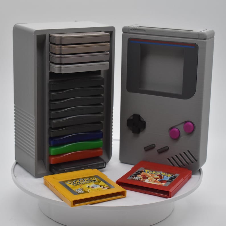以下が含まれることがあります： グレーの3Dプリントされたゲームボーイカートリッジホルダーとゲームボーイのレプリカ。ホルダーには、さまざまな色のゲームカートリッジがいくつか入っています。2つのゲームカートリッジが前にあり、1つは黄色、もう1つは赤です。ゲームボーイのレプリカにはピンク色のボタンがあります。