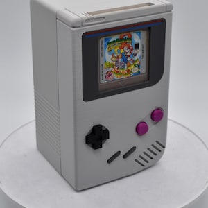 以下が含まれることがあります： スーパーマリオランドのゲームカートリッジを表示する画面を備えた、グレーの携帯型ゲーム機。コンソールには黒い十字キー、ピンクのボタン、赤いアクセントが付いています。ゲームカートリッジには「DMG-MD-USA」と「Made in Japan」と記載されています。