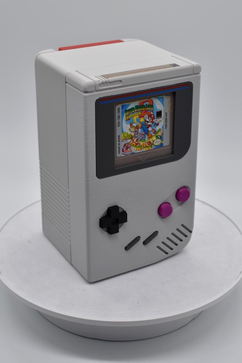 以下が含まれることがあります： スーパーマリオランドのゲームカートリッジを表示する画面を備えた、グレーの携帯型ゲーム機。コンソールには黒い十字キー、ピンクのボタン、赤いアクセントが付いています。ゲームカートリッジには「DMG-MD-USA」と「Made in Japan」と記載されています。
